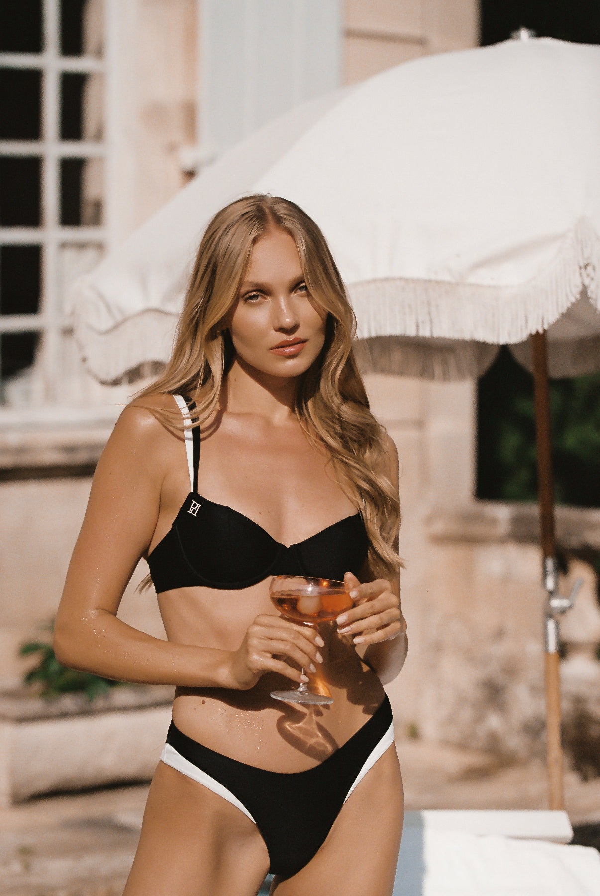Monaco black ivory bikini made of European swim fabric and 24 kt gold-plated hardware, back side view of model holding a drink against a chateau and umbrella backdrop.