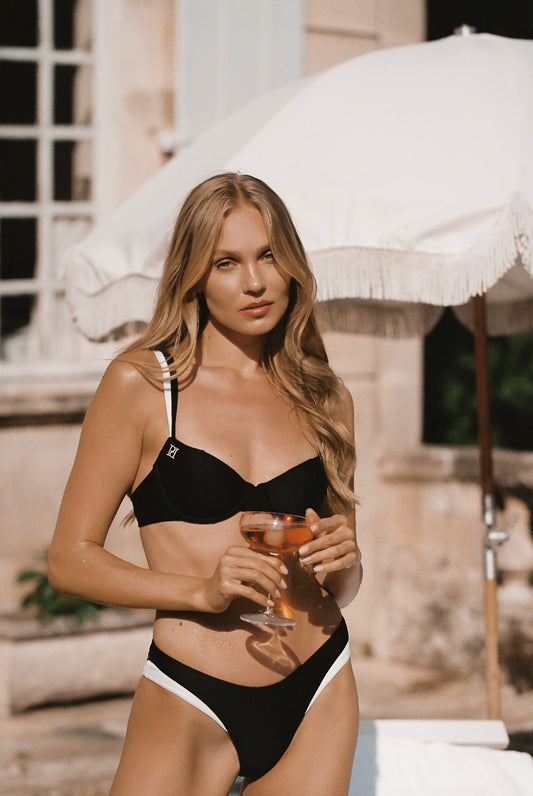 Monaco black ivory bikini made of European swim fabric and 24 kt gold-plated hardware, back side view of model holding a drink against a chateau and umbrella backdrop.